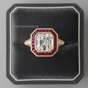May include: A gold ring featuring a large, square, clear central stone, encircled by a halo of small, red gemstones. The ring is presented in a dark grey velvet box. The text "sierrajewelsca" is visible.