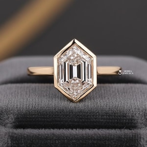 May include: A gold ring with a hexagonal-shaped diamond. The diamond is set in a bezel setting and has a clear, faceted appearance. The ring is displayed on a gray velvet surface. The ring is a piece of jewelry.