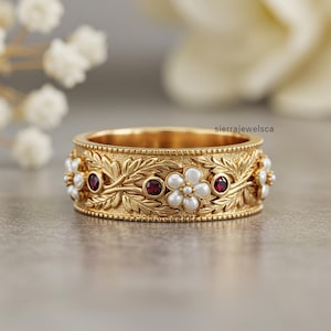 Puede incluir: Un anillo dorado con un diseño floral. El anillo presenta detalles de flores blancas y piedras preciosas rojas, junto con un patrón de hojas detallado. El anillo es una toma de primer plano, mostrando los intrincados detalles.