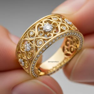 Può includere: Un anello in filigrana dorata con un design traforato dettagliato. L'anello presenta diverse pietre preziose rotonde e chiare e una fascia bordata con pietre più piccole. L'anello è tenuto tra due dita.