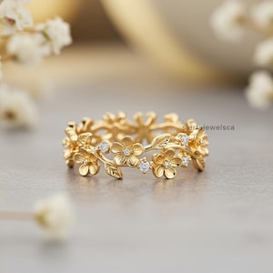 Op de afbeelding: Een gouden ring met een doorlopende band van gevormde bloemen en bladeren met kleine ingelegde heldere stenen. Het metaal heeft een gepolijste afwerking en de tekst sierrajewelsca is zichtbaar aan de rechterkant van het ontwerp.