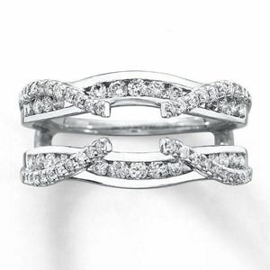 May include: A silver ring guard with a curved design. The ring guard features two intertwined bands, each encrusted with small, sparkling diamonds. The bands curve around the center, creating a unique and elegant look. The ring guard is designed to enhance the appearance of a ring.