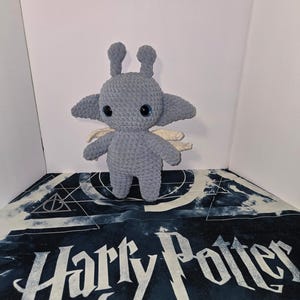 Könnte beinhalten: Ein handgefertigtes, graues gehäkeltes Wesen mit großen schwarzen Augen, kleinen Flügeln und zwei Antennen. Das Plüschtier steht auf einem Harry-Potter-Stoff mit den Worten "Harry Potter" in Weiß.
