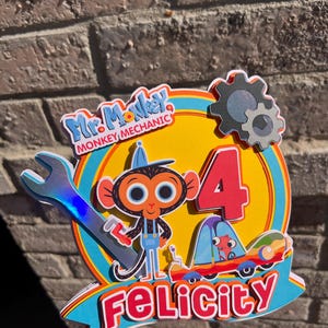 May include: A colorful cake topper featuring Mr. Monkey, a cartoon monkey mechanic, with a wrench and a red car. The topper also includes the number 4 and the name Felicity.