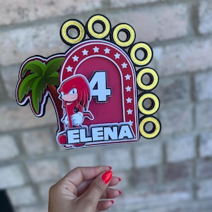 May include: Red and black cake topper featuring a cartoon character with a palm tree and gold rings. The number 4 and the name Elena are also included.