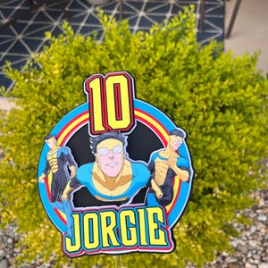 May include: A colorful cake topper featuring a cartoon character with the number "10" in a red and yellow font. The name "JORGIE" is displayed in yellow block letters. The design includes blue, yellow, and red accents, perfect for a birthday celebration.
