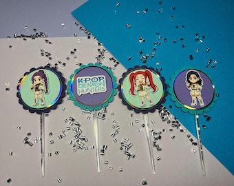 May include: Four cupcake toppers with cartoon illustrations of K-Pop idols. Each topper has a scalloped edge and a clear stick. One topper has the text "K-POP DEMON HUNTERS". The background is white and blue with silver confetti.