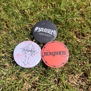 May include: Three round buttons with the text "MAYHEM" and "LVDA GAGA" in white on a black button, "THE MAYHEM BALL" in red on a red button, and "THE MAYHEM BALL" with a cross design on a white button.