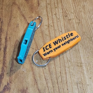 May include: Two keychains on a wooden surface. One is a teal whistle, and the other is an orange tag with the text "ICE Whistle Warn your neighbors!". Both have silver key rings.