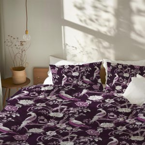 May include: A bed with a dark purple duvet cover and pillowcases featuring a white and lavender bird and floral pattern. The bedding is on a wooden bed frame with white pillows. The room has a neutral color palette with natural light.