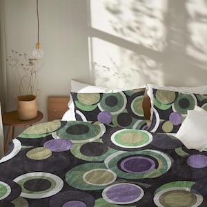 May include: Bedding set featuring a duvet cover and pillowcases with a retro-style design. The pattern includes overlapping circles in shades of green, purple, and white against a dark background. The bedding is on a bed with white sheets.