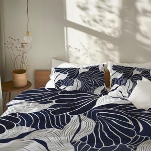 May include: A navy and white floral bedding set with a large-scale leaf design. The duvet cover and pillowcases feature a navy blue background with white leaf-like patterns. The bed is made with white sheets and pillows. A small wooden side table is visible.