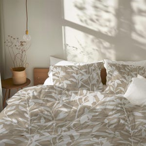 May include: A bed with a beige and white floral patterned duvet cover and pillowcases. The bedding is on a wooden bed frame. A small side table with a woven basket and dried flowers is next to the bed. A light fixture hangs above the bed.