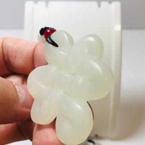 Puede incluir: Un colgante de jade blanco con forma de flor con una cuenta roja en un cord&oacute;n negro.