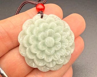 May include: A jade pendant carved in the shape of a flower with a light green color. The pendant is attached to a brown cord with a red bead.