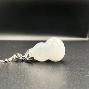 May include: A white gourd-shaped pendant necklace with a silver chain. The pendant is a light, opaque color and has a smooth, rounded shape. The necklace is displayed against a dark background.