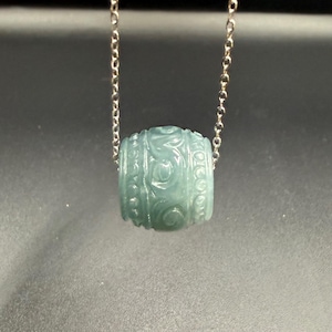 Peut inclure: Un collier avec pendentif en jade vert clair. Le pendentif est en forme de baril avec des motifs floraux sculptés. La chaîne du collier est argentée et délicate. Le fond est un dégradé de gris.