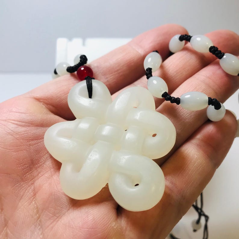Puede incluir: Collar con colgante de jade blanco con un dise&ntilde;o anudado y un cord&oacute;n negro con cuentas blancas. El colgante tiene forma de flor con cinco p&eacute;talos.