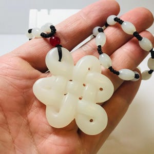 Puede incluir: Collar de jade blanco con un colgante de nudo y un cord&oacute;n negro. El colgante tiene forma de flor con un agujero en el centro. El collar est&aacute; hecho de cuentas de jade blanco y un cord&oacute;n negro.