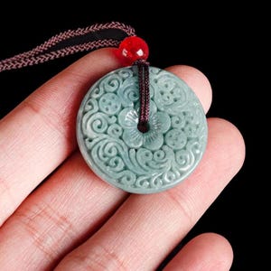 Peut inclure: Un collier avec un pendentif en jade rond de couleur verte orné de sculptures florales complexes. Le pendentif est attaché à un cordon brun avec une perle rouge.