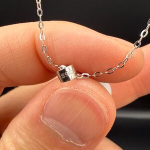 May include: Close-up of a delicate silver necklace. The chain is made of small, interconnected links. A small, rectangular clasp with the marking "S925" is visible. The necklace is held against a blurred background.