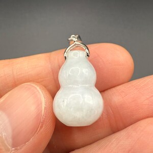 May include: A small, white gourd-shaped pendant with a silver-colored bail. The pendant is smooth and translucent, with subtle variations in color. The bail is a simple, curved design. The pendant is held in a person's hand.