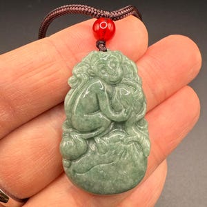May include: A green jade pendant carved with a monkey holding a peach, a symbol of longevity and good fortune in Chinese culture. The pendant is attached to a brown cord with a red bead.