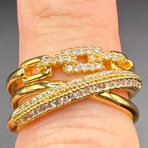 Op de afbeelding: Gouden ring met drie banden, elk versierd met heldere zirkonia-stenen. De bovenste band heeft een vierkant ontwerp met een kettingschakelpatroon.