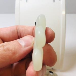 Puede incluir: Un colgante de jade blanco tallado en forma de flor con cinco p&eacute;talos. El colgante es liso y pulido, con un brillo sutil.