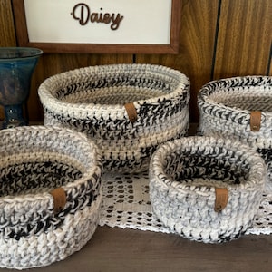May include: Set of four crocheted baskets in black and white with a gray and white ombre effect. The baskets are nested inside each other and have a brown leather tag.