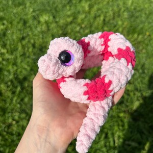 May include: A handmade crochet snake toy in shades of pink and red. The plush snake has large, glittery eyes and a soft, textured appearance. The toy is held in a hand against a background of green grass.