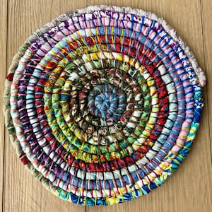 May include: A round, colorful trivet made from fabric scraps. The trivet features a spiral design with a variety of colors including red, blue, green, and yellow. The trivet is approximately 8 inches in diameter and is suitable for placing hot dishes.