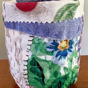 May include: A handmade fabric container with a patchwork design. The container features a variety of floral patterns in green, blue, purple, and red, with a blue band and white trim. The stitching is visible, adding to the handcrafted aesthetic.