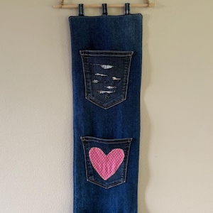 May include: A denim wall hanging with three pockets. The top pocket has a distressed look. The middle pocket has a pink heart with white polka dots. The bottom pocket has a red floral design.