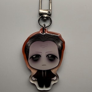 May include: A keychain featuring a cartoon illustration of a man in a black suit with a white shirt and black tie. The keychain has a silver clasp and a clear acrylic body with an orange border.