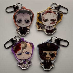 May include: Four acrylic keychains featuring cartoon illustrations of horror movie characters. Each keychain has a silver-colored clasp. The characters are depicted in various outfits and with different facial expressions, with a focus on their distinctive features.