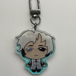 May include: A keychain featuring a cartoon character with white hair and a detailed outfit. The character is set against a light blue background. The keychain has a silver-colored clasp.