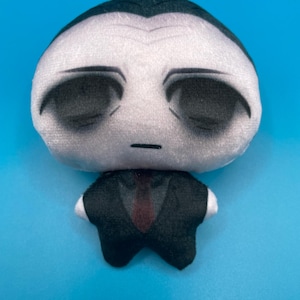 May include: A plush doll with a pale face, black eyes, and a small mouth. The doll is wearing a black suit with a red tie and white cuffs. The doll has a small white tuft of hair on top of its head.
