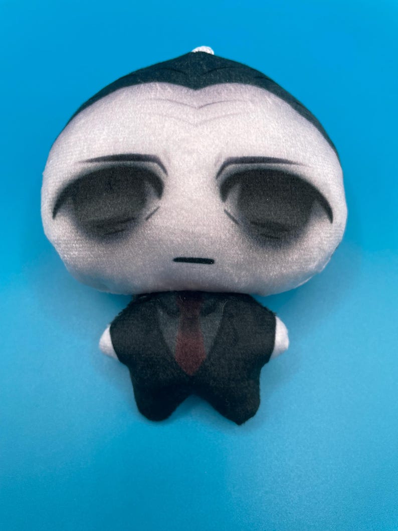May include: A plush doll with a pale face, black eyes, and a small mouth. The doll is wearing a black suit with a red tie and white cuffs. The doll has a small white tuft of hair on top of its head.