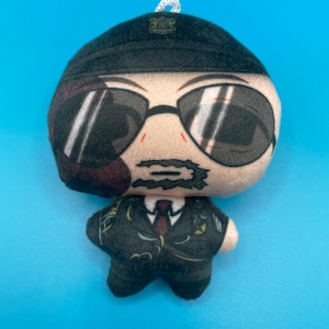 May include: A plush doll in the shape of a person wearing a black suit, sunglasses, and a black cap. The doll has a white string loop at the top for hanging. The background is a solid light blue.