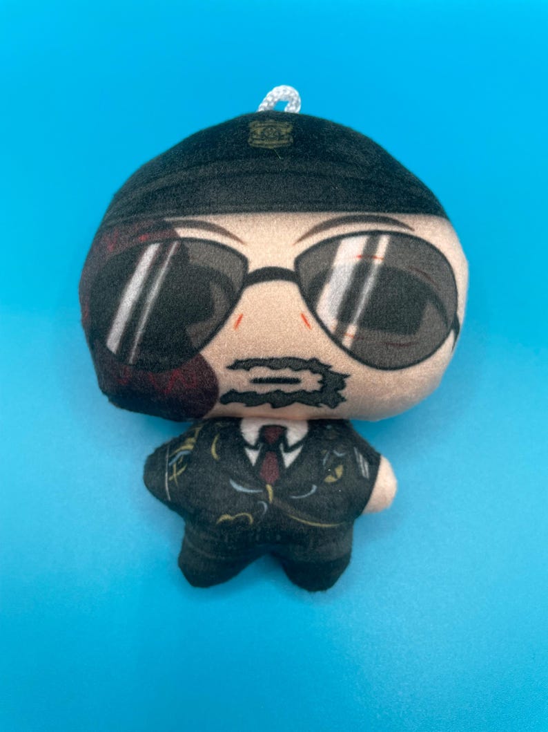 May include: A plush doll in the shape of a person wearing a black suit, sunglasses, and a black cap. The doll has a white string loop at the top for hanging. The background is a solid light blue.