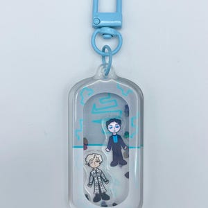 May include: A clear acrylic keychain with a light blue clasp and ring. Inside the keychain are two small character illustrations and light blue decorative accents. The characters are wearing detailed outfits. The background is a light gray.
