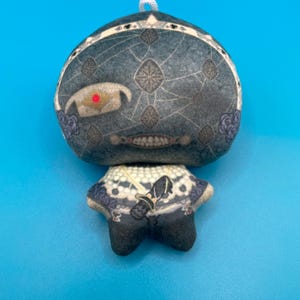 May include: A plush doll with a round head and a small body. The head is dark gray with a geometric pattern and a single red eye. The doll has a white and blue patterned collar and black feet. A white string is attached to the top of the head.