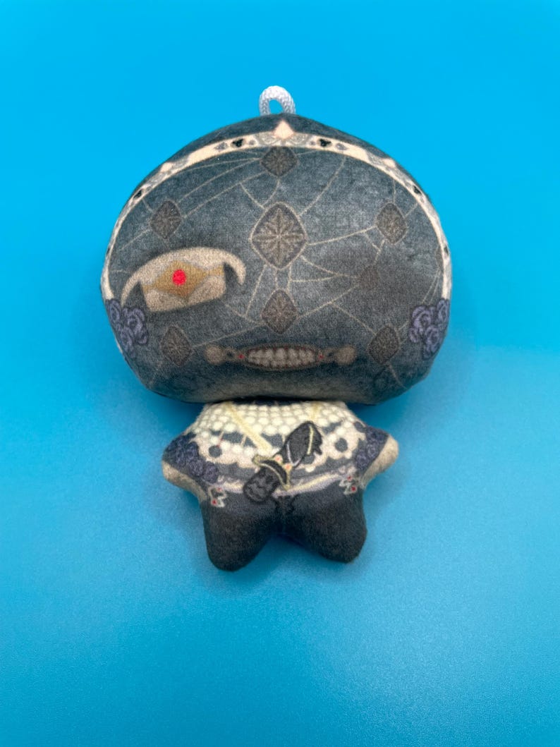 May include: A plush doll with a round head and a small body. The head is dark gray with a geometric pattern and a single red eye. The doll has a white and blue patterned collar and black feet. A white string is attached to the top of the head.