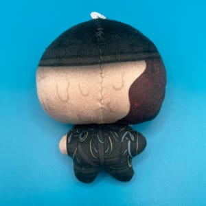 May include: A plush doll with a black and tan head, and a black outfit with yellow accents. The doll has a white string at the top of its head and is set against a blue background.