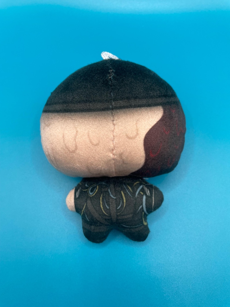 May include: A plush doll with a black and tan head, and a black outfit with yellow accents. The doll has a white string at the top of its head and is set against a blue background.