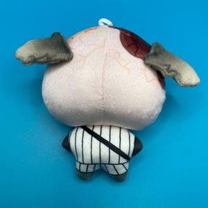 May include: A plush toy shaped like an eye with small, gray, wing-like appendages. The eye is light pink with brown and red markings. The lower portion of the toy is a white and black striped design with gray feet.