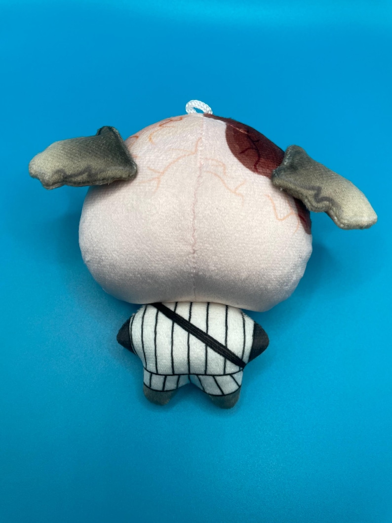 May include: A plush toy shaped like an eye with small, gray, wing-like appendages. The eye is light pink with brown and red markings. The lower portion of the toy is a white and black striped design with gray feet.