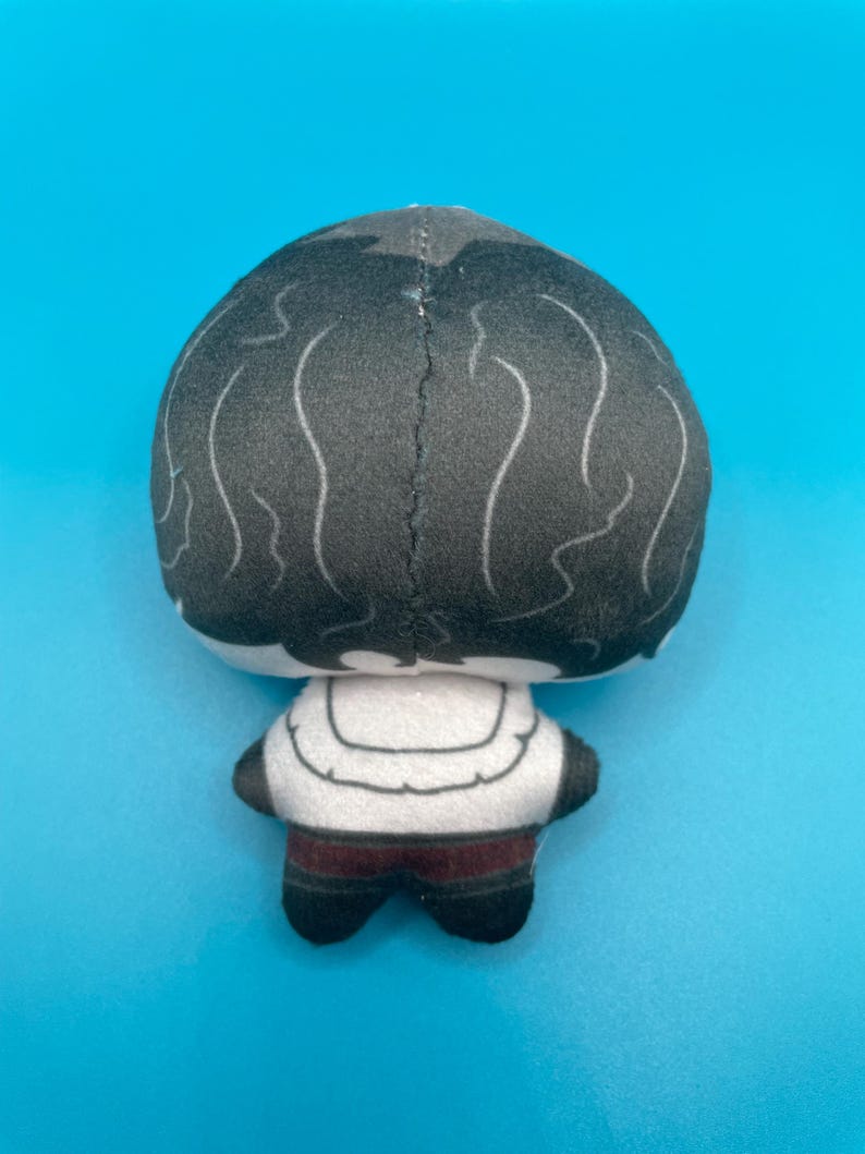 May include: A plush doll with a black head and white wavy lines, a white torso with a curved design, and dark red pants. The doll's back is visible against a bright blue background.