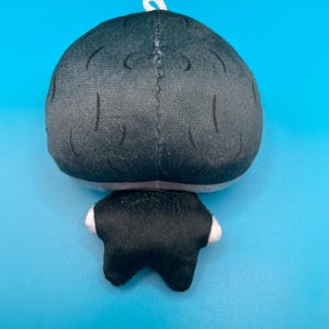 May include: A plush doll with a black head and body, and white accents on the feet. The doll has a white loop at the top for hanging. The background is a solid light blue.